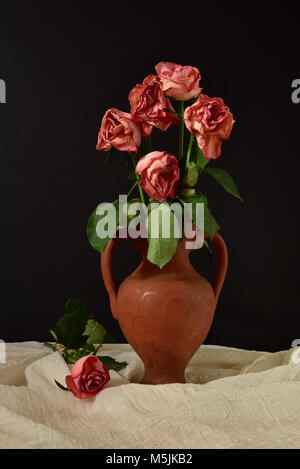 Cinq roses roses mourir dans un vase en terre cuite sur un fond noir, avec une seule rose sur tissu de mousseline. Banque D'Images