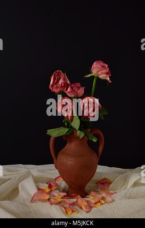 Cinq roses roses mourir dans un vase en terre cuite, sur un tissu de mousseline parsemé de pétales, sur un fond noir. Banque D'Images