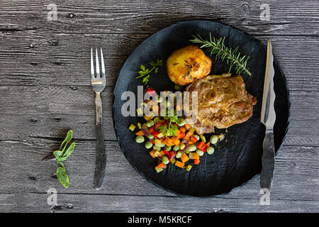 Porc grillé avec des légumes grillés et une pomme de terre au four sur une plaque noire Banque D'Images