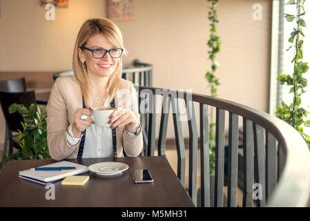 Young business woman sitting in coffee shop à table en bois, boire du café et prendre des notes. Sur la table sont pen, réserver et téléphone mobile. Banque D'Images