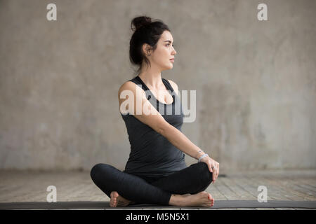 Young woman practicing yoga, faisant tourné exercice facile, Parivrtta Sukhasana posent, de travail, le port des vêtements de sport, pantalon noir et haut, indo Banque D'Images