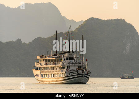 Un bateau de pêche traditionnel vietnamien squd passe derrière un grand bateau de croisière touristique au coucher du soleil sur la baie d'Halong, Vietnam Banque D'Images