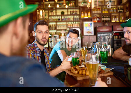 Groupe de jeunes hommes discutant avec animation les uns avec les autres et la consommation de boissons alcoolisées pendant que traîner dans pub moderne, certains d'entre eux portant ha vert Banque D'Images