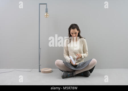 Belle fille lit un livre tout en s'assied sur le plancher dans lotus poser à côté de la lampe longue avec edison ampoule sur un fond de mur gris en studio. Elle Banque D'Images