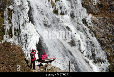 25 février 2018, l'Allemagne, Todtnau : excursionnistes s'asseoir sur un banc regardant la gelée en partie Todtnauer de cascades. Le Todtnauer cascade dans la région de la Forêt-Noire du sud est l'un des plus hauts cascades naturelles en Allemagne. Photo : Steffen Schmidt/dpa Banque D'Images