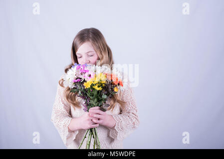 12-year-old fille avec de longs cheveux blond sale, holding bouquet de marguerites colorées sur la face inférieure et sentir avec les yeux fermés, fond blanc Banque D'Images