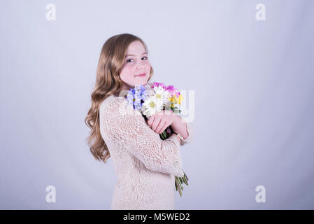 Smiling 12-year-old fille avec de longs cheveux blonds, sales et incliné d'un bouquet de marguerites colorées serrant contre fond blanc Banque D'Images