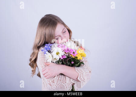Smiling 12-year-old fille avec de longs cheveux blond sale, serrant le bouquet de marguerites colorées avec les yeux fermés contre fond blanc Banque D'Images