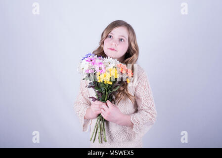 12-year-old fille avec de longs cheveux blond sale, holding bouquet de marguerites colorées tout en regardant vers le haut et vers le côté contre fond blanc Banque D'Images