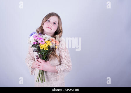 12-year-old fille avec de longs cheveux blond sale, holding bouquet de marguerites colorées tout en regardant vers le haut et vers le côté contre fond blanc Banque D'Images
