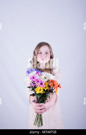 Smiling 12-year-old fille avec de longs cheveux blond sale, holding out bouquet de marguerites colorées à contre fond blanc ; fille de se concentrer Banque D'Images