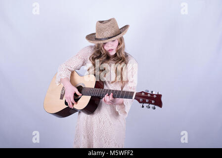 12-year-old fille avec de longs cheveux blonds, sales et wearing straw hat playing acoustic guitare à six cordes against white background Banque D'Images