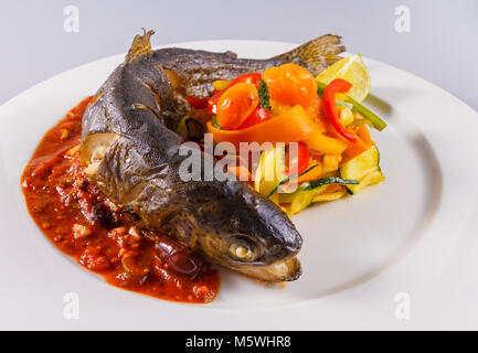 Truite grillée entière avec légumes rôtis et sauce tomate servie sur une élégante assiette de restaurant. Banque D'Images