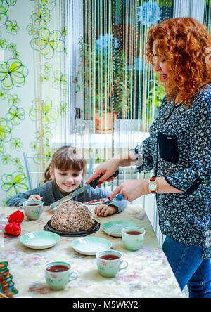 La petite fille est assise à la table avec une cuillère dans sa main et attend que sa maman pour lui couper un morceau de délicieux gâteaux. Banque D'Images