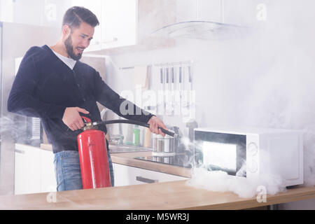 Jeune homme pulvérisant extincteur sur le four à micro-ondes dans la cuisine Banque D'Images