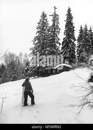 Un snowboard girl à un casque bleu se distingue avec sa main sur le côté d'un conseil orange vif dans la forêt et ressemble à une vieille maison de bois h Banque D'Images