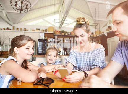 Famille heureuse avec deux filles en train de dîner au restaurant du smartphone et l'utilisation. La famille, la parentalité, la technologie concept. Banque D'Images