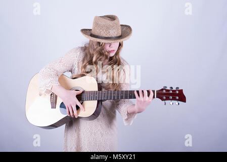 12-year-old fille avec de longs cheveux blonds, sales et wearing straw hat couvrant les yeux playing acoustic guitare à six cordes against white background Banque D'Images