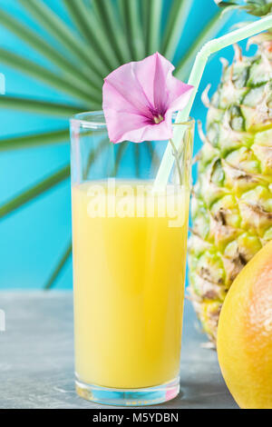 Composition de Grand Verre à jus de fruits tropicaux fraîchement pressé avec de la paille et l'ensemble de la fleur de pamplemousse ananas. Feuilles de palmiers sur fond bleu Banque D'Images