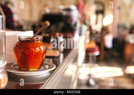 Miel savoureux dans un bocal en verre avec une cuillère en bois au premier plan dans un café avec un arrière-plan flou. Banque D'Images