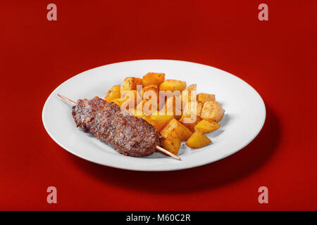 Kebab de viande de porc, de boeuf avec pommes de terre sur une plaque blanche. Contexte monophonique rouge. Un repas copieux, le dîner. Pour le menu. Vue de côté Banque D'Images