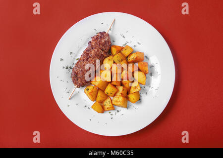 Kebab de viande de porc, de boeuf avec pommes de terre sur une plaque blanche. Contexte monophonique rouge. Un repas copieux, le dîner. Pour le menu. Vue de dessus Banque D'Images