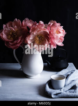 Coral Charm de pivoines en pleine floraison, dans un pot sur le tableau blanc avec tasse et théière, tetsublin fond sombre, tourné en lumière naturelle Banque D'Images