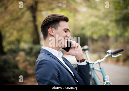 Young attractive businessman talking on smartphone à l'extérieur dans un parc, avec son vélo à côté de lui Banque D'Images