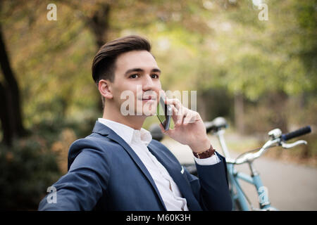 Young attractive businessman talking on smartphone à l'extérieur dans un parc, avec son vélo à côté de lui Banque D'Images