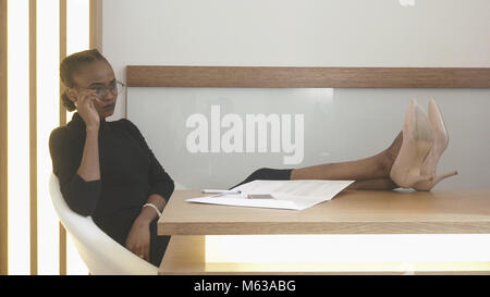 Portrait de la charmante femme africaine avec natural make-up à la réception. La pensive brunette prend les lunettes off alors qu'elle repose avec les jambes sur la table. L'emplacement du bureau. Banque D'Images