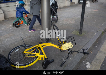 Un dockless Ofo tombé de vélo loué se trouve sur le trottoir comme un jeune enfant scoots passé avec sa mère à Herne Hill, le 26 février 2018, dans le sud de Londres, en Angleterre. Banque D'Images