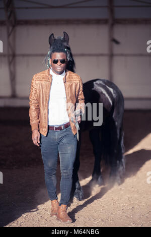 African Man wearing sunglasses près de black horse in hangar Banque D'Images