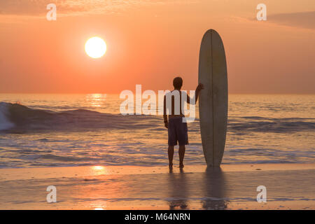 Surfer avec longboard debout sur plage au coucher du soleil en regardant les vagues Banque D'Images