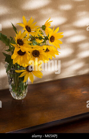 Les tournesols jaune vif dans un vase en cristal assis sur une table en bois, avec du soleil à travers feuilles à venir contre un mur d'or Banque D'Images