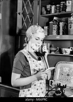 1930 Femme ménagère en tablier, debout dans la cuisine à parler au téléphone - T4522 HAR001 HARS CHERS nostalgie à l'intérieur de 20 à 25 ans 25 à 30 ANS AU FOYER AU FOYER DU BONHEUR LA LIBERTÉ DE CHOIX DES FEMMES AU FOYER DU SERVICE À LA CLIENTÈLE CONNEXION Téléphones Téléphones MID-ADULT MID-ADULT WOMAN WOMAN B&W NOIR ET BLANC PORTRAIT DE L'ORIGINE ETHNIQUE DES PERSONNES À L'ANCIENNE Banque D'Images