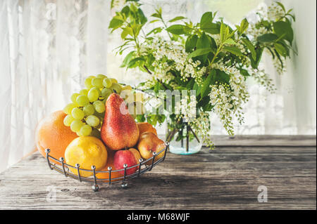 Fruit poire, pamplemousse, citron, pêche sont dans le panier sur l'arrière-plan d'un bouquet de petites fleurs blanches de bird cherry sur une table rustique sur une Sun Banque D'Images