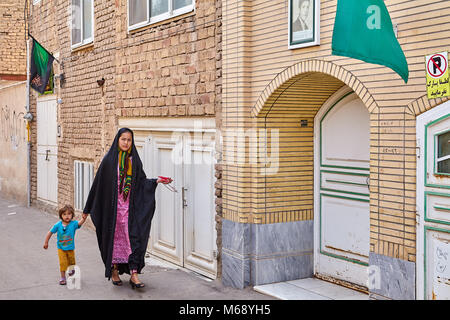 Kashan, Iran - avril 27, 2017 : Une jeune femme musulmane, dans un tchador noir, jeté sur une robe de couleur, les promenades le long de la rue avec une petite fille, qui Banque D'Images