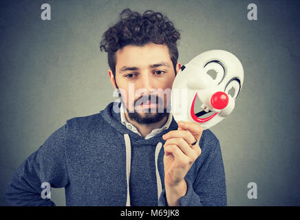 Jeune homme bouleversé barbu portant sur la personnalité de happy clown masque sur fond gris. Banque D'Images