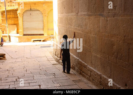 Silhouette noire d'un petit garçon marche à travers une arche dans une rue ensoleillée, balayant d'une main le long du mur en pierre massive. Rajastan, Inde. Banque D'Images