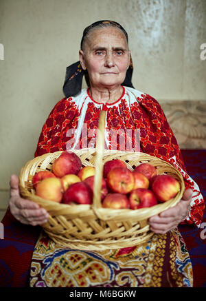 Vieux fermier femme en costume traditionnel tenant un panier plein de pommes Banque D'Images