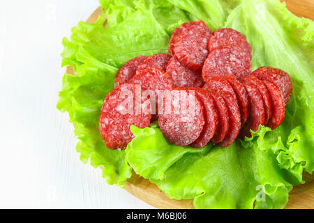 Saucisse fumée, salami coupé en tranches sur une salade sur une circulaire en bois à découper sur un tableau blanc Banque D'Images