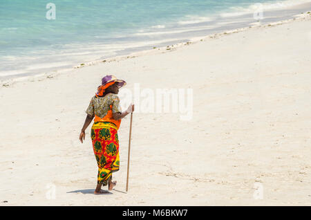 Zanzibar, Tanzanie - le 4 décembre 2014 : Femme avec des vêtements colorés et une cabane à marcher le long de la plage de Nungwi. Banque D'Images