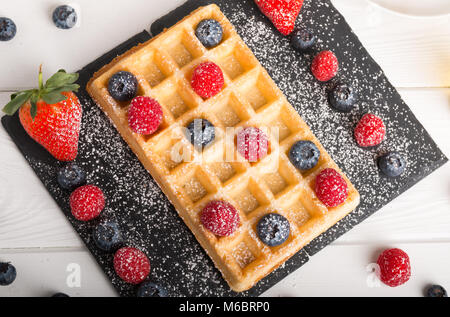 Gaufres de Bruxelles fraîchement cuits sur une plaque en pierre noire avec des fraises fraîches et de bleuet et de framboise est sur une table en bois blanc avec du miel et c Banque D'Images