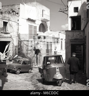 Années 1950, historiques, l'heure d'été et une petite rue étroite avec des véhicules italiens de l'époque, une petite bulle 'fiat' et un 'spaCar', à trois roues d'un camion de livraison, un Piaggio Ape 400, stationné sur l'inégale de galets, sur l'île méditerranéenne de l'île d'Elbe, Toscane, Italie, Banque D'Images