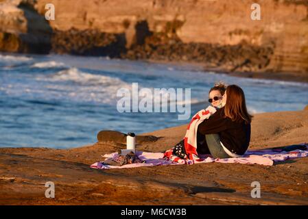 Deux jeunes femmes dans le contrat cadre un pique-nique au coucher du soleil le long de falaises d'or avec le fracas des vagues de l'océan au coucher du soleil les falaises, San Diego, Californie, USA. Banque D'Images