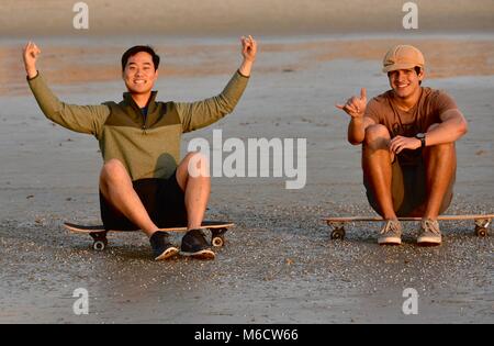 La hanche, deux jeunes hommes assis sur une planche à roulettes regardant le coucher du soleil sur la plage, ce qui rend 'haka' signe (chill out), à San Diego, Californie, USA Banque D'Images