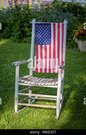 Vieux rustique peint blanc rocking chair assis dans cour avec drapeau américain drapé sur l'arrière Banque D'Images