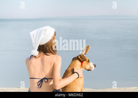 Jeune personne de sexe féminin à santa claus hat s'asseoir sur la plage à côté de staffordshire terier chiots avec toy reindeer horns Banque D'Images