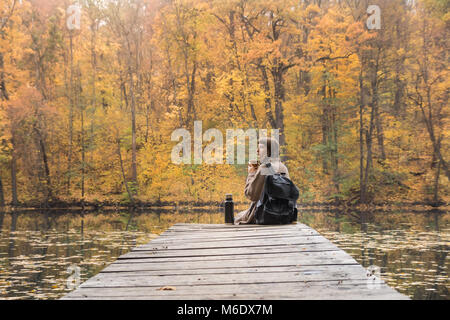 Young female hiker with backpack se trouve au rivage, boit du café et ressemble à de beaux paysages de l'été indien Banque D'Images