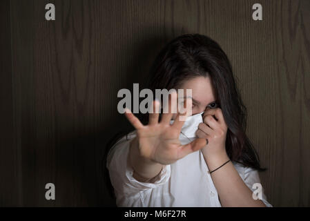 Jeune femme cachant son visage avec sa chemise tout en étendant la main vers l'appareil photo afin de ne pas être vu, sur un fond de bois, dans les ombres. Banque D'Images